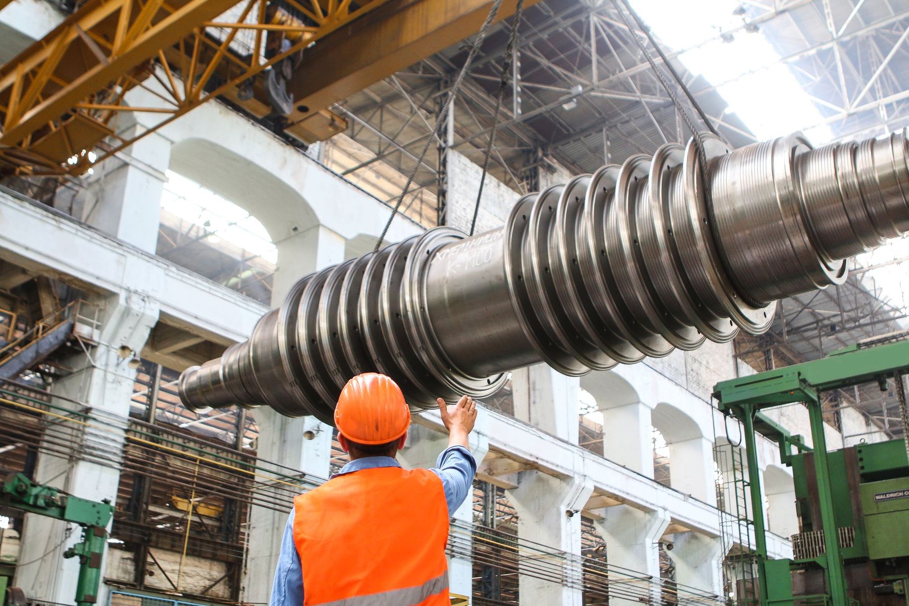 A factory floor with a large machined shaft suspended from an overhead crane